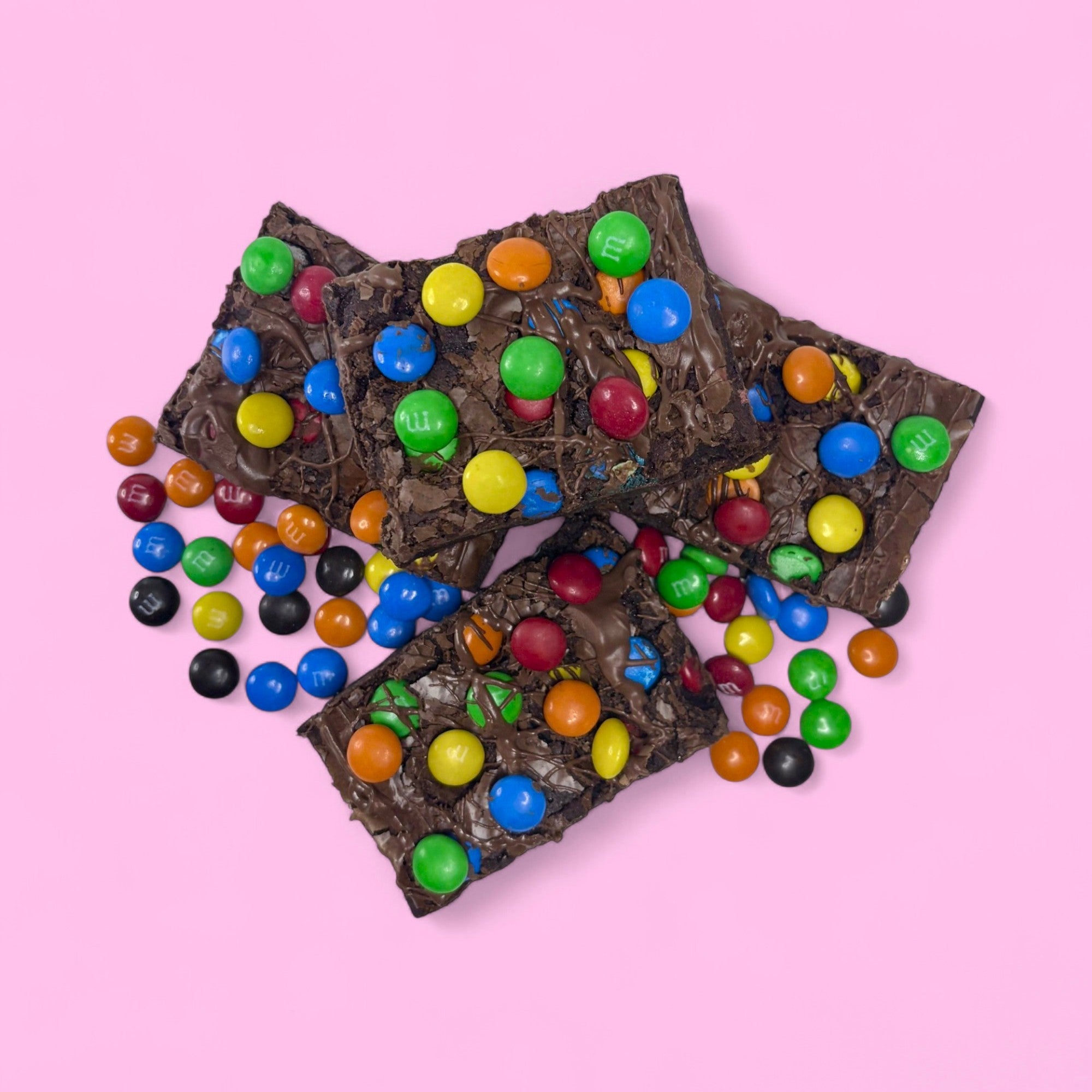 four m & m brownies stacked surrounded by an array of colourful milk chocolate m & ms, made with real belgian chocolate sitting against a pink background