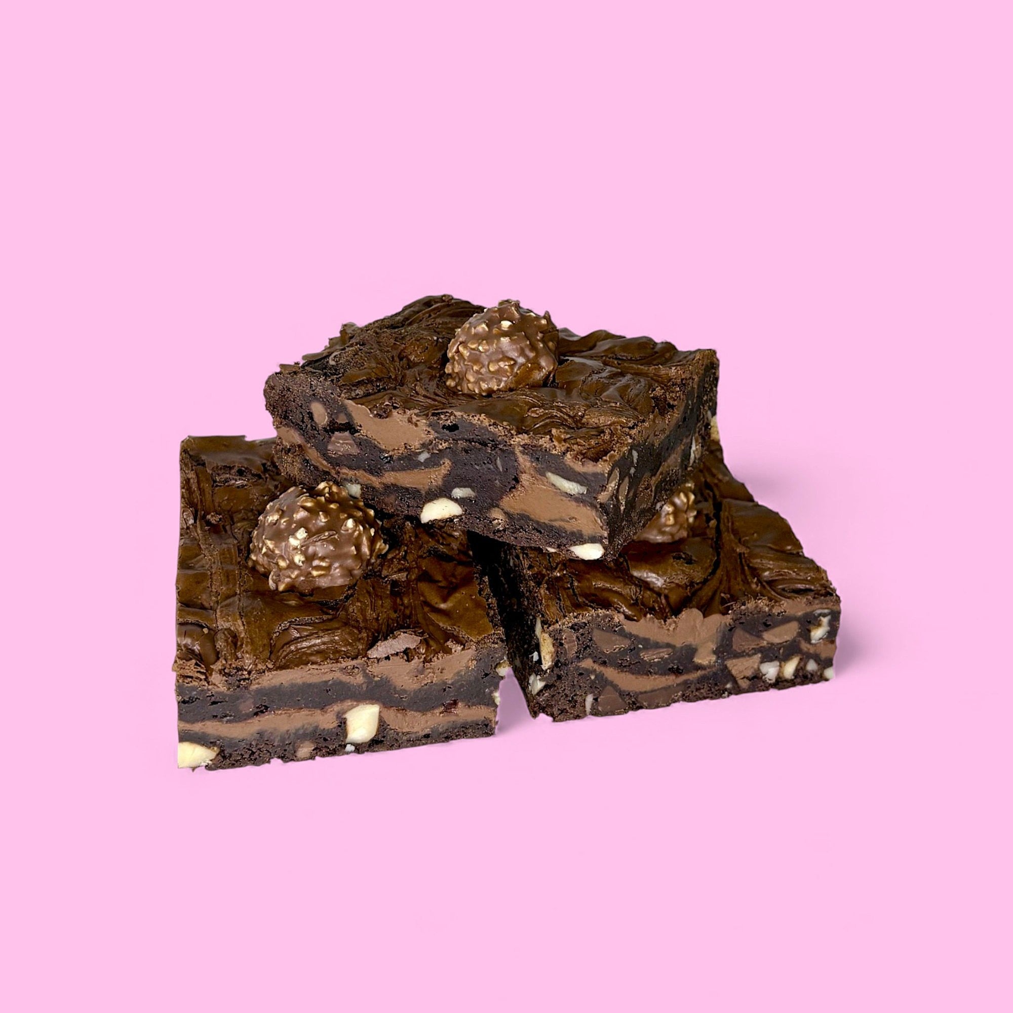three nutella fudge brownies made with real belgian chocolate, featuring roasted hazelnuts inside, layers of nutella and ferrero rochers on top sitting against a pink background