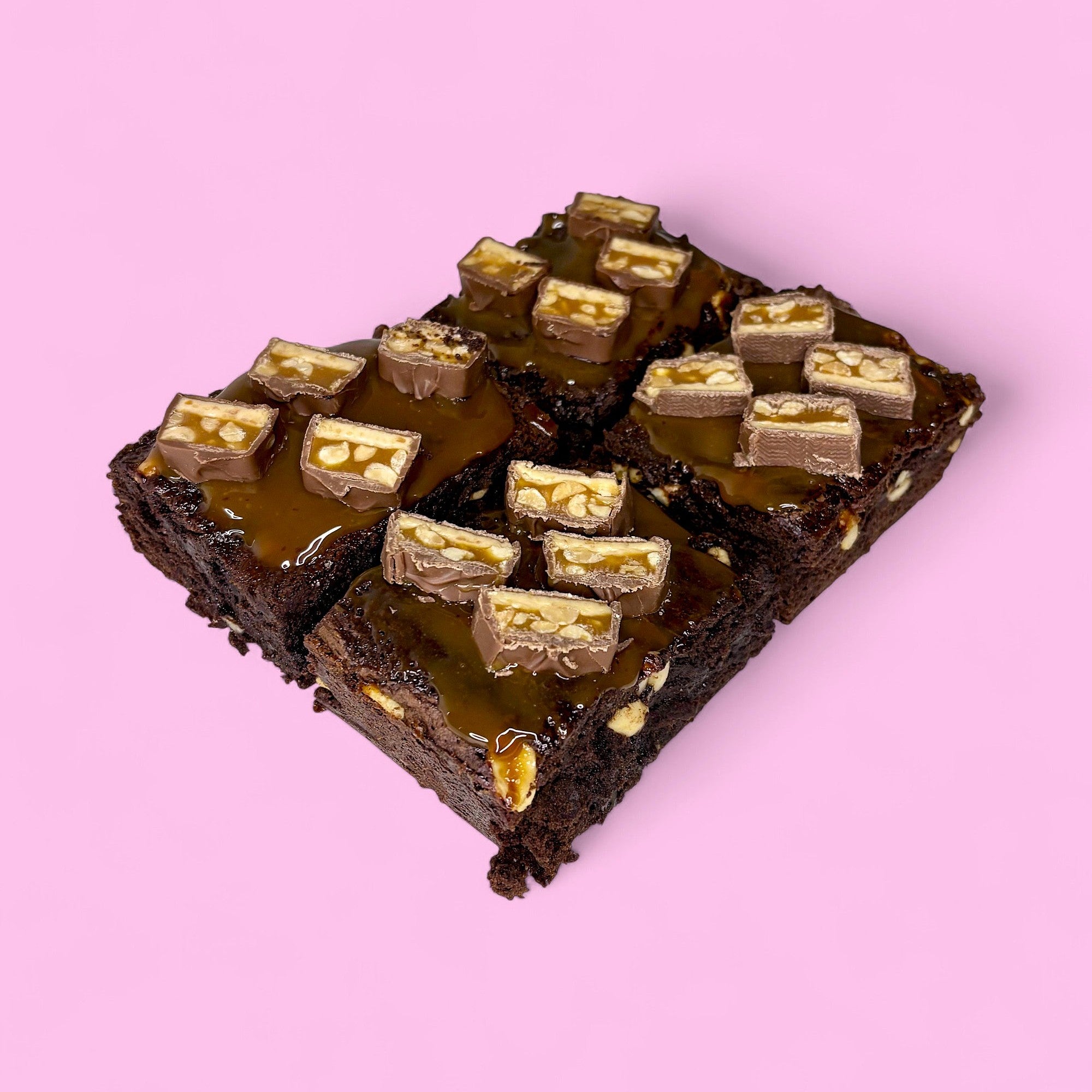 Chocolate fudgy brownies filled with crunchy peanuts and smothered with salted caramel, made with real belgian chocolate and sitting against a pink background