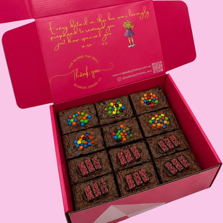 Box of 12 brownies with colorful toppings on a pink background in open hot pink gift box