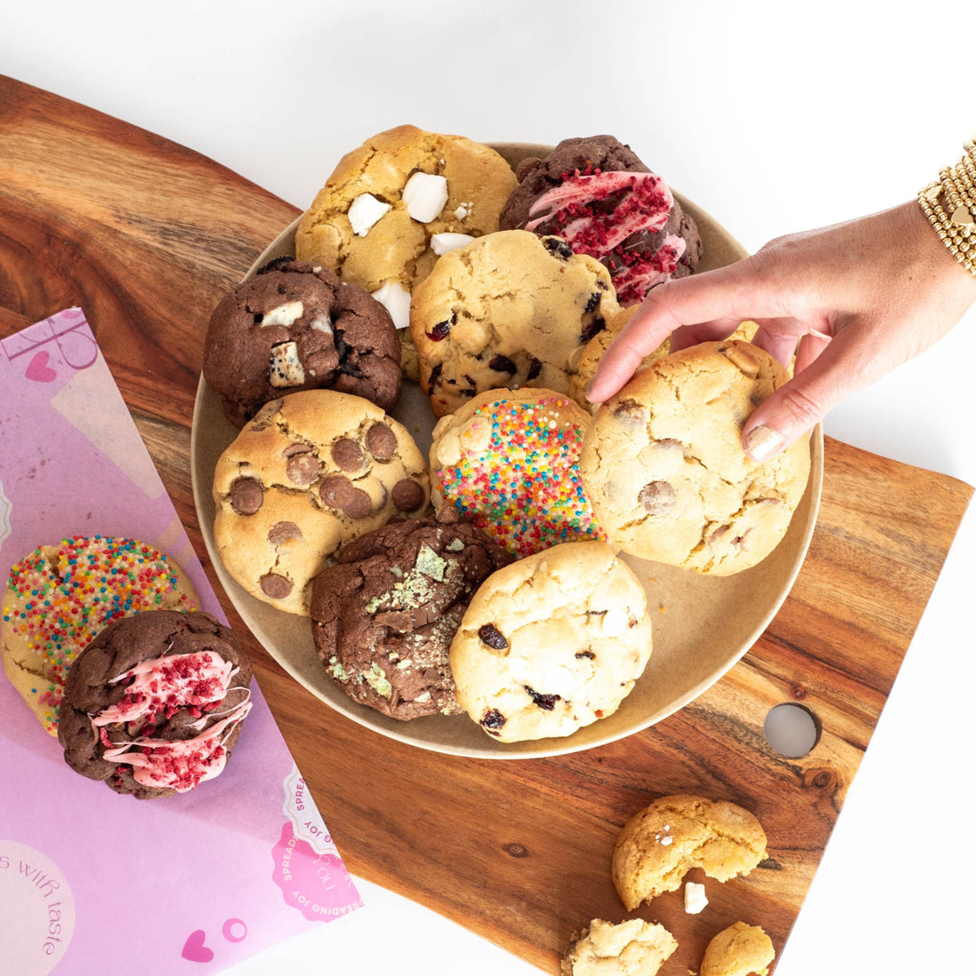A plate of assorted chunky cookies on a chopping board with a hand reaching in and taking one cookie