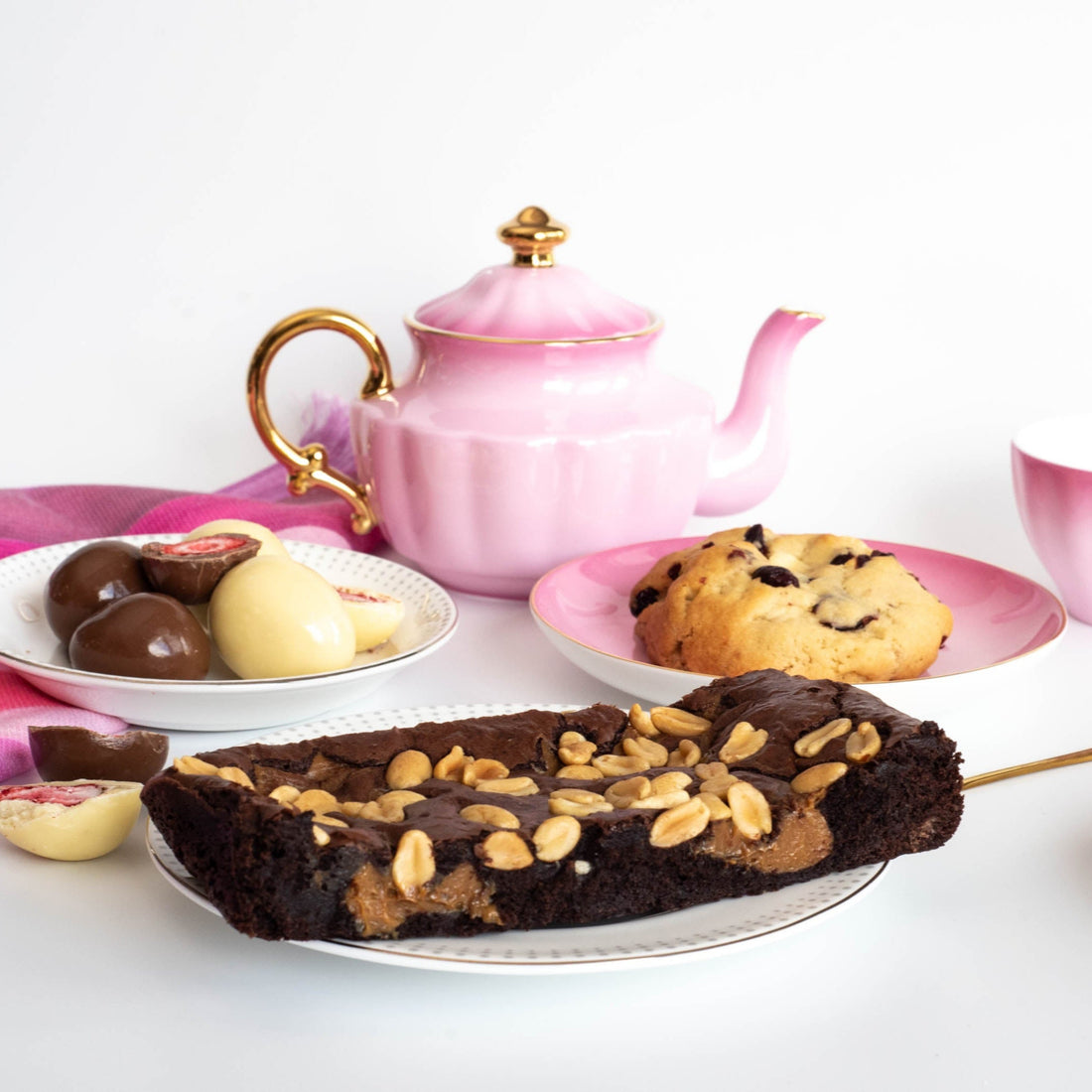 assorted desserts including a brownie slab, a single cookie and cut chocolate covered strawberries sit on pretty plates in front of a pink teapot