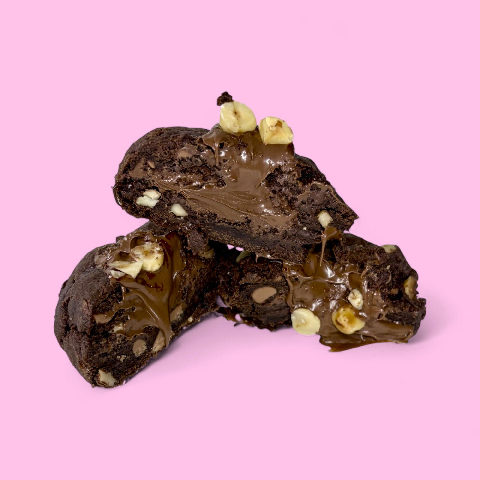 Chocolate chunky cookies filled with nutella and roasted hazelnuts, made with real belgian chocolate, sitting against a pink background