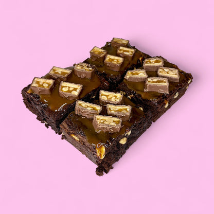 Chocolate fudgy brownies filled with crunchy peanuts and smothered with salted caramel, made with real belgian chocolate and sitting against a pink background