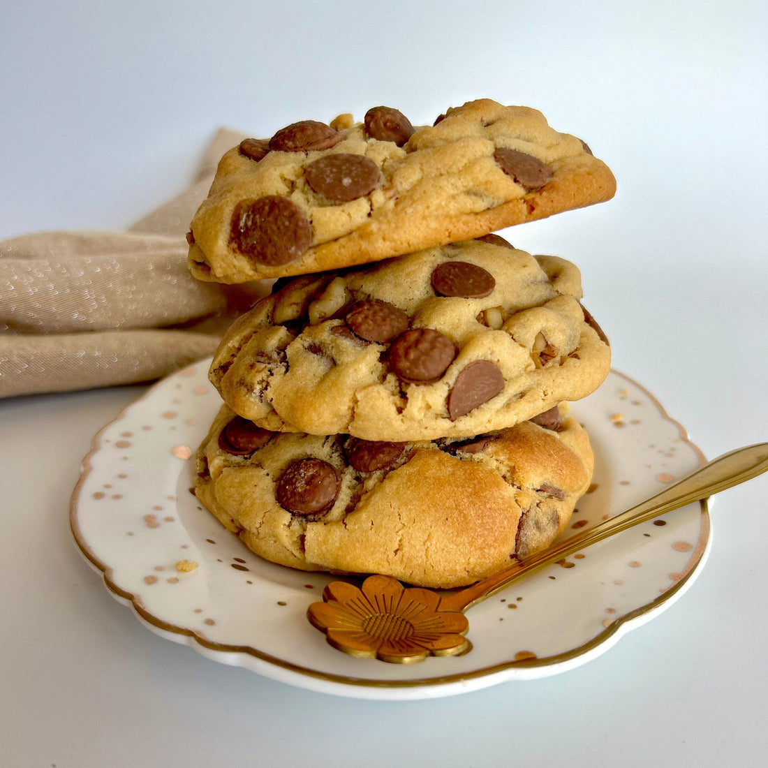 Three choc chip walnut cookies stacked on top of each other sitting on a white and gold plate with a gold spoon
