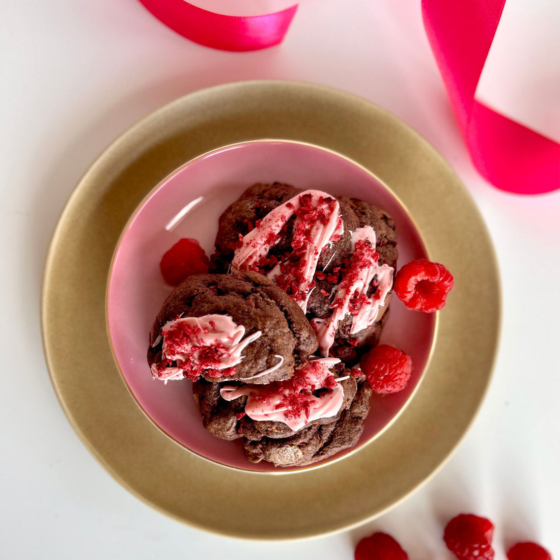 birds eye view of choc raspberry chunky cookies with chocolate dough, pink icing and raspberries scattered on top sitting on a pink plate