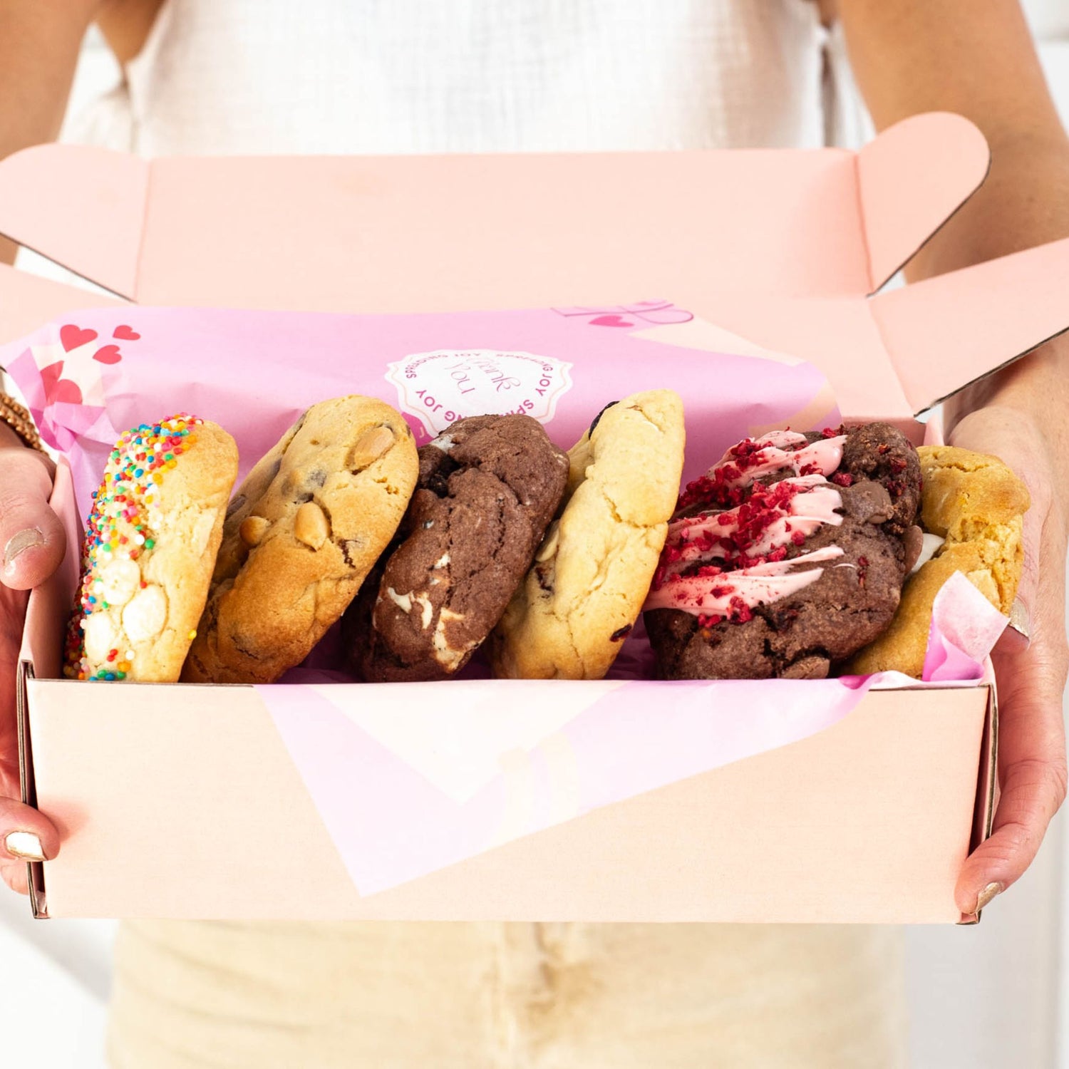 hands holding a pink gift box with pretty packaging and 6 yummy cookies on their side lined up ready to be indulged