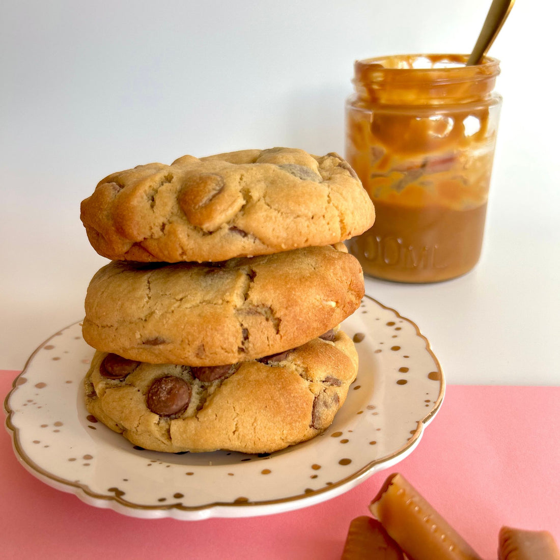 three freshly baked cookies stacked on top of each other with choc chips and caramel in a glass container in the background. pink and white background