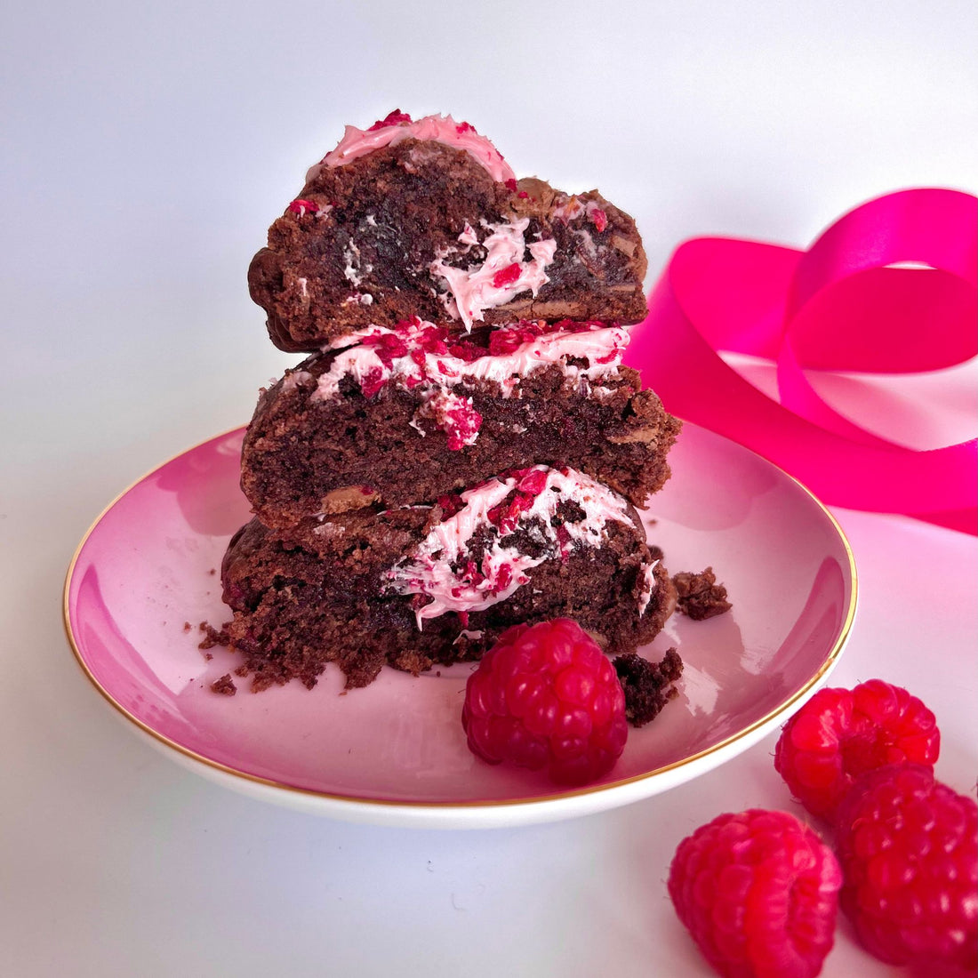 Cookies chopped in half sitting on a pink plate with pink ribbon in background. Cookies are chocolate dough with raspberry pieces throughout