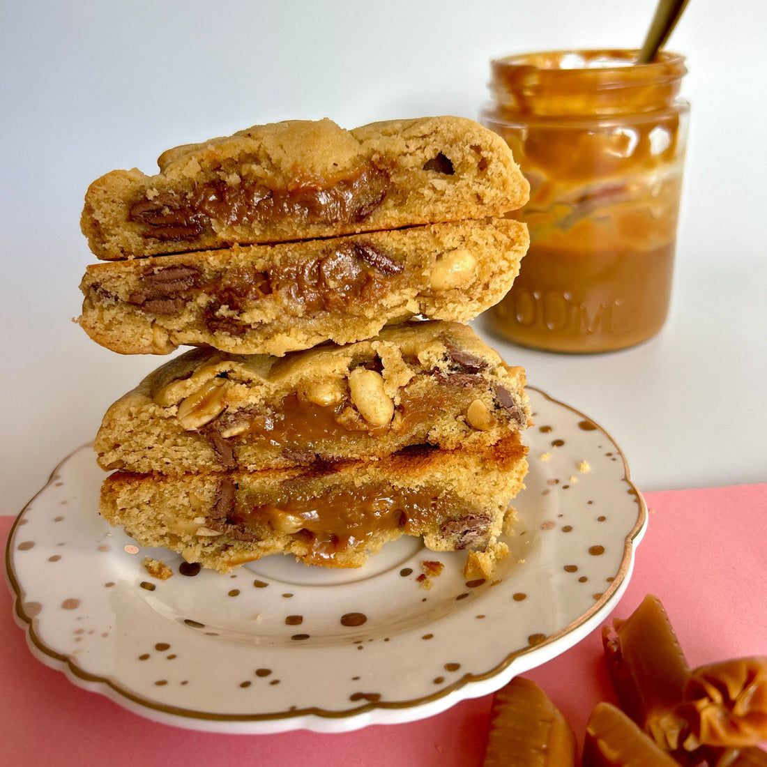 two peanut caramel cookies cut in half and stacked on top of each other on a white and gold plate, the middle of the cookie shows gooey caramel and golden peanuts with a jar of caramel in the background