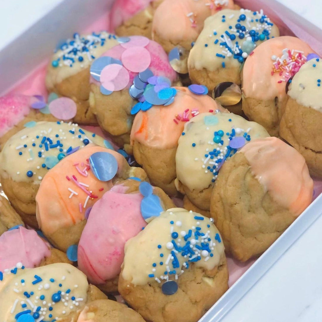 A gift box displayed with mini party birthday cookies in pink, yellow and orange colored icing with confetti